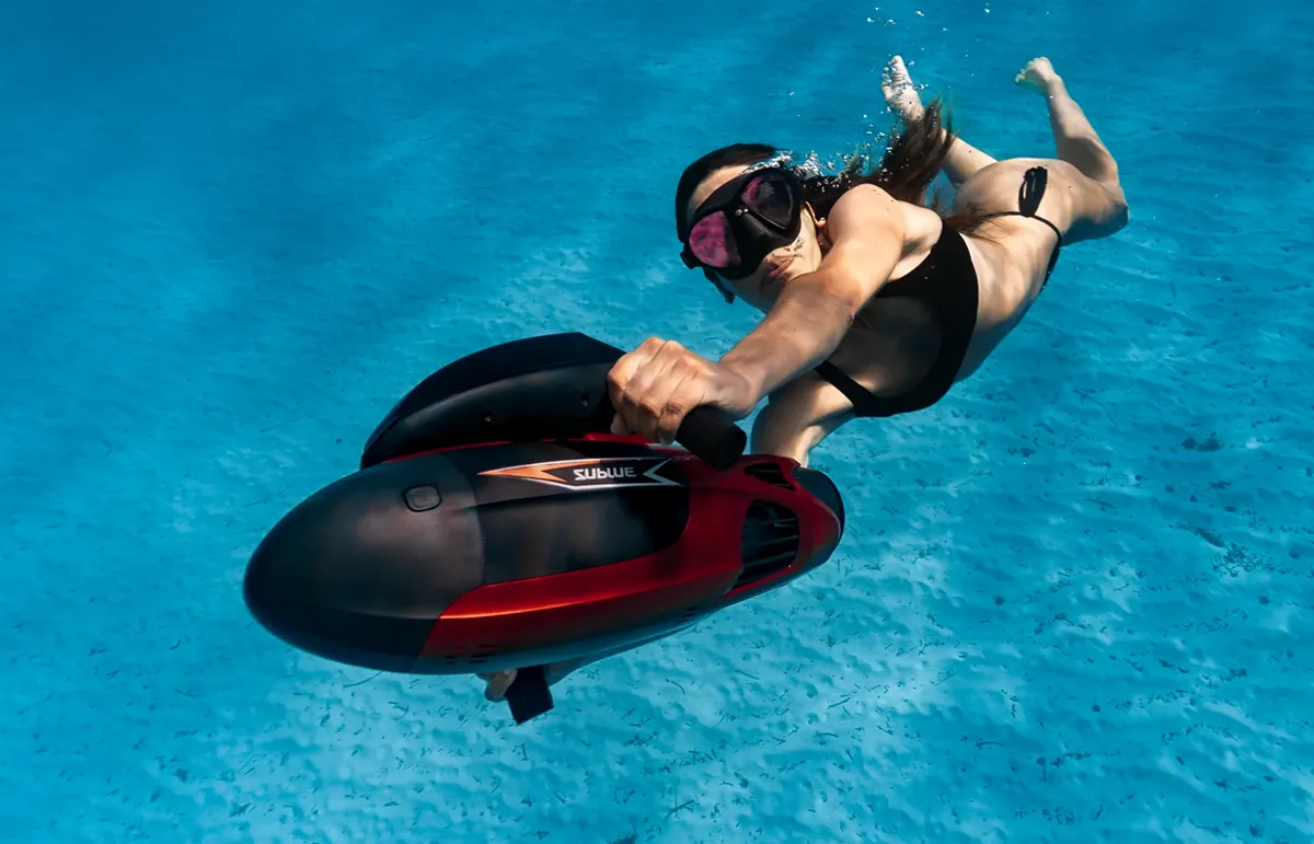 Woman in a black bikini wearing a snorkeling mask rides a red and black underwater scooter in clear blue water.