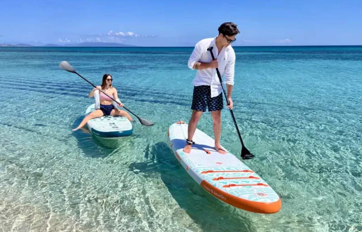 A man stands on a paddleboard with a paddle in crystal-clear turquoise water, while a woman sits on another board nearby under a sunny sky.
