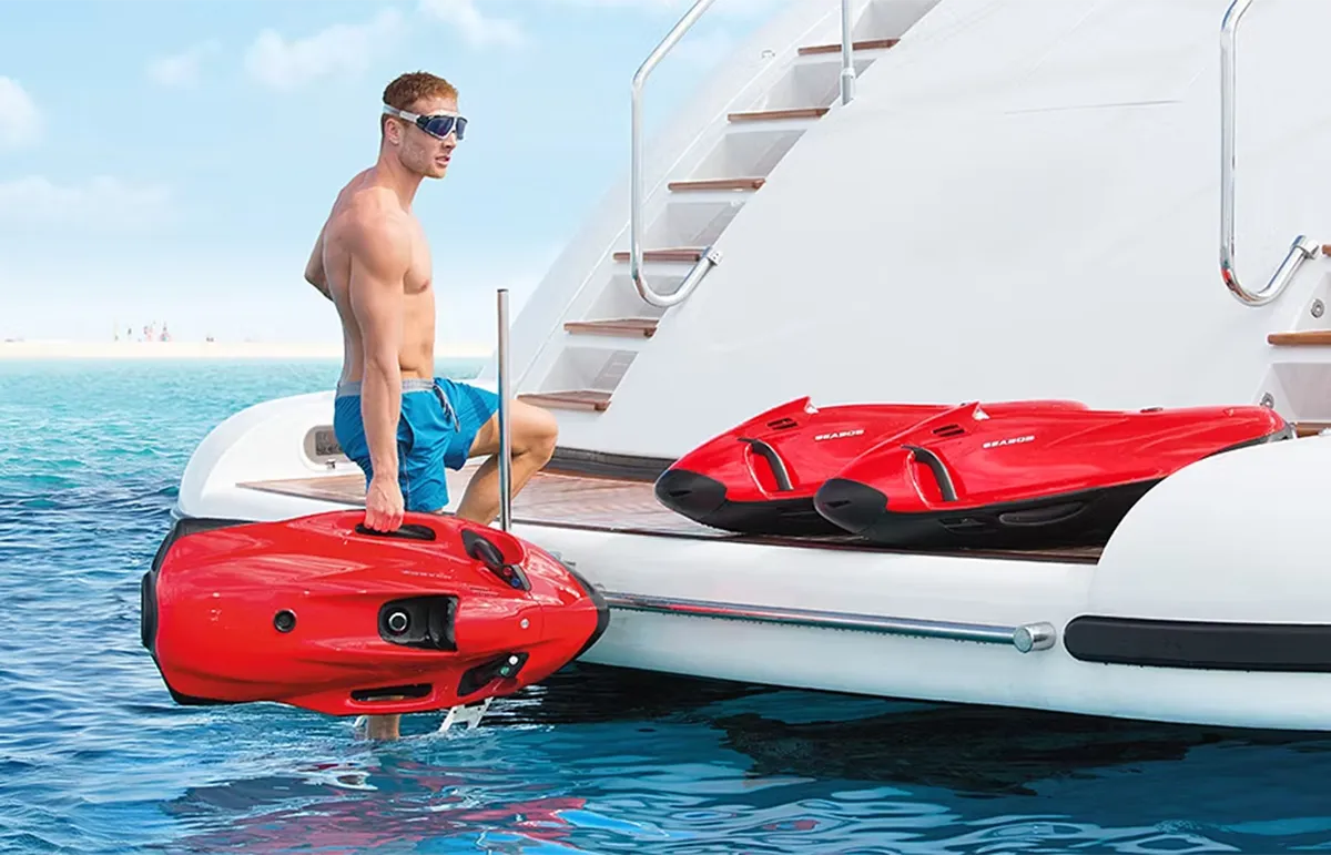 Man in blue swim trunks and sunglasses steps onto a red inflatable boat from a white yacht, with two red jet skis on deck.