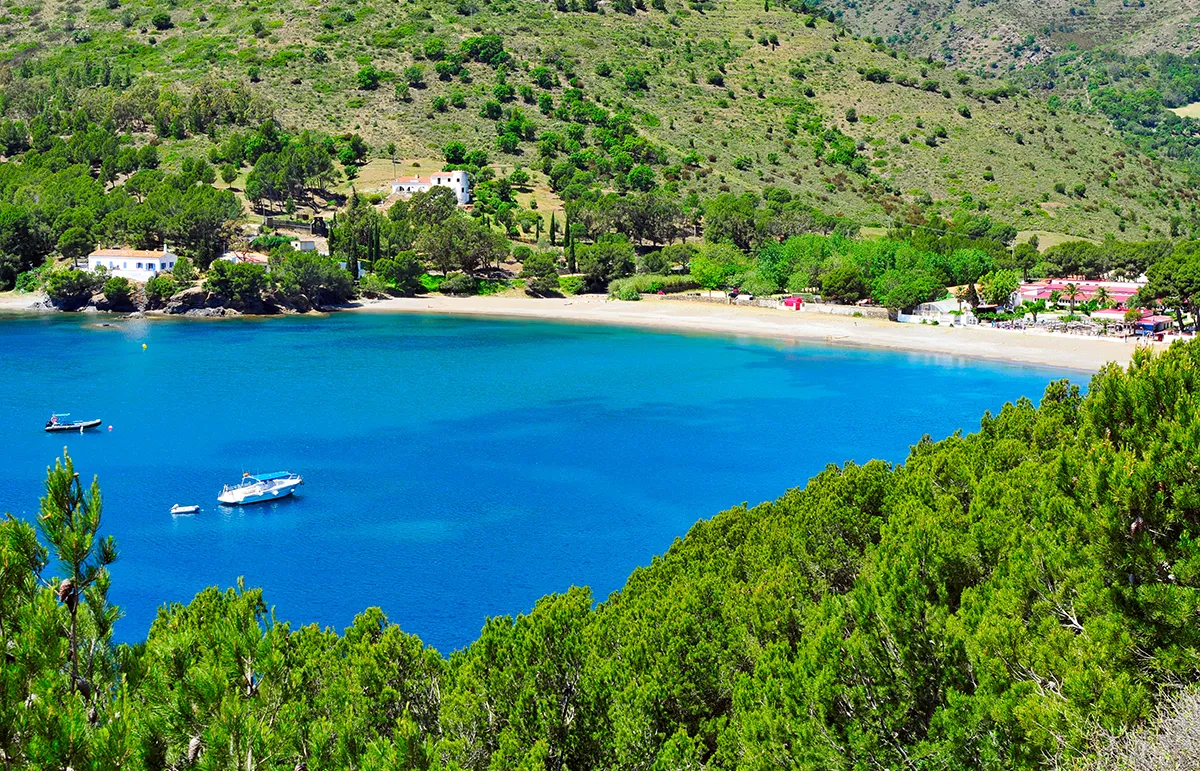 Scenic coastal cove with turquoise water, boats near a sandy beach and green hillside village in the background.