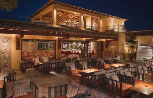 Nighttime outdoor lounge with multiple seating areas, wooden tables, patterned cushions, and candle lighting.