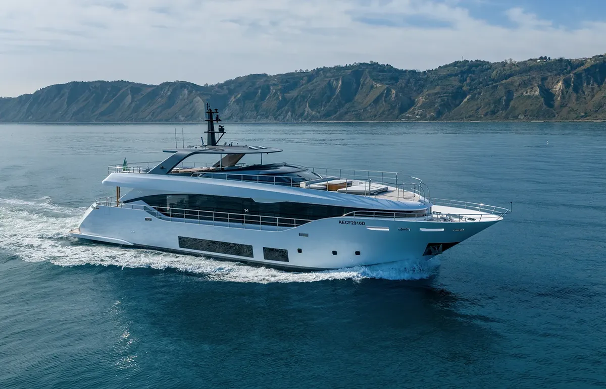 White luxury motor yacht sailing on calm blue sea with green hills in the distance.