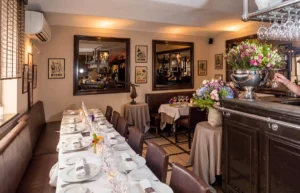 Long dining table set for a formal meal in a cozy restaurant, with white tablecloths, wine glasses, candles, and floral centerpieces, beige walls and mirrors in the background.
