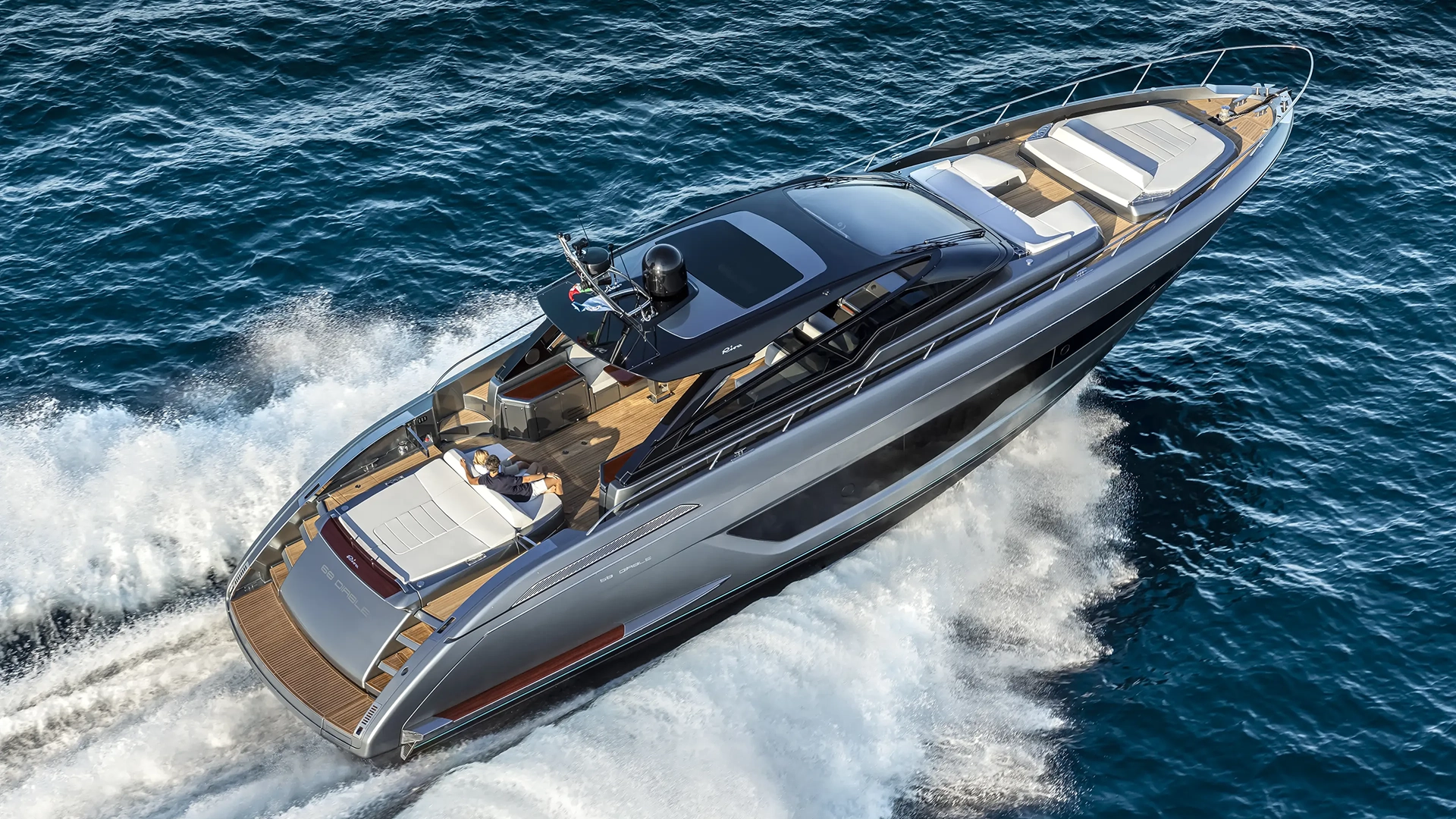 giocava-riva68-diable1920x1080px10 - Echonautique.com Sleek luxury motor yacht cutting through blue ocean with white wake behind it. Two people lounge on the rear deck as the boat speeds forward.